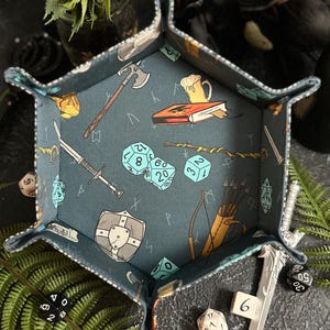 May include: A blue and gray hexagonal dice tray with a fantasy theme. The tray features a sword, axe, book, dice, and other fantasy imagery.