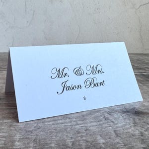 Może przedstawiać: Biała, złożona wizyt&oacute;wka stołowa z czarnym nadrukiem Mr. & Mrs. Jason Burt oraz cyfrą 8 wyśrodkowaną pod nazwiskiem. Kartka jest ustawiona w formie namiotu, eksponując elegancką czcionkę pisankową na przednim panelu wizyt&oacute;wki.
