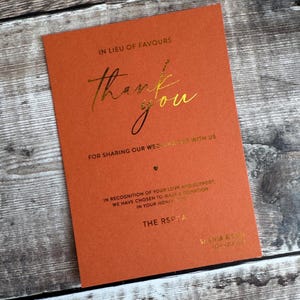 Pode incluir: Cart&atilde;o laranja com texto dourado. O cart&atilde;o diz "Thank you" e &eacute; para um casamento. O texto diz "Em vez de lembran&ccedil;as" e "Por partilhar o nosso dia de casamento connosco". Tamb&eacute;m menciona uma doa&ccedil;&atilde;o.