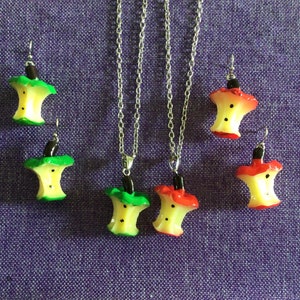 May include: A set of three necklaces and earrings featuring a bitten apple design. The apples are yellow with red and green accents. The necklaces are silver chains.