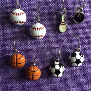 May include: A set of 7 pairs of sport-themed earrings. The earrings are silver with a variety of sports balls and a bowling pin. The balls include a baseball, basketball, football, and a soccer ball.