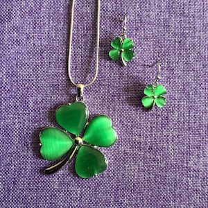 Op de afbeelding: Een zilveren ketting en oorbellenset met groene vierbladige klavertjes charms. De klavertjes charms hebben een glanzende, iriserende afwerking.
