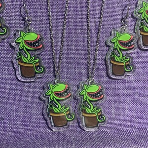以下が含まれることがあります： ネックレスとイヤリングを含む、植物をテーマにしたジュエリーのコレクション。各作品は、茶色の鉢に入った赤い裏地のある口を持つ緑色の植物を特徴としています。ジュエリーは透明なアクリル製で、銀色のチェーンとイヤリングフックから吊り下げられています。