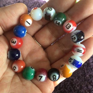 Puede incluir: Un círculo de 15 canicas de vidrio de colores con los números del 1 al 15. Las canicas están dispuestas en círculo y se sostienen en una mano.