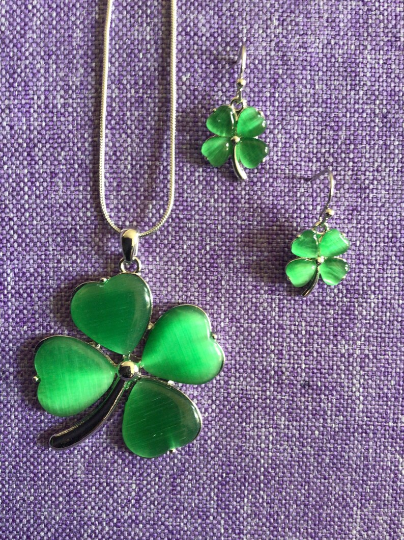 May include: A silver necklace and earrings set featuring green four-leaf clover charms. The clovers have a shiny, iridescent finish.