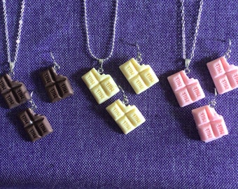 May include: A collection of chocolate bar-shaped jewelry, including necklaces and earrings. The chocolate bars are in shades of brown, white, and pink, with the word "MILK" visible on each piece. The jewelry hangs against a purple fabric background.