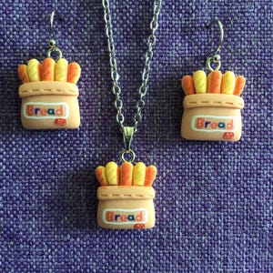 May include: A jewelry set featuring a necklace and earrings. The set includes a silver chain necklace with a pendant and a pair of earrings. Each piece has a charm shaped like a bag of bread, with yellow and orange bread pieces and the word "Bread".