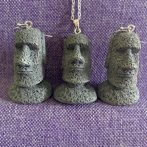 May include: A jewelry set consisting of two gray, textured Moai head earrings and a matching pendant on a silver-toned chain. Each piece features the distinct facial features of a stone statue with a porous, rough surface texture and neutral gray color.