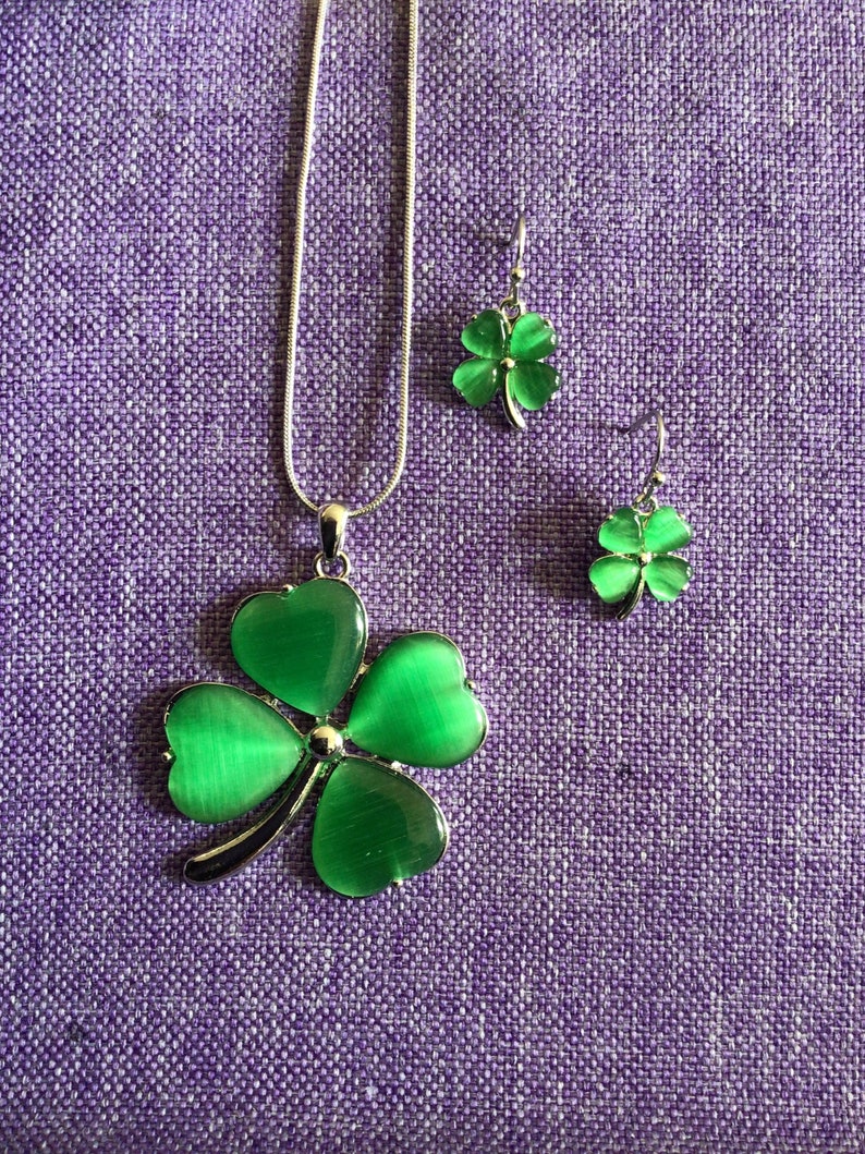 May include: A silver necklace and earrings set featuring green four-leaf clover charms. The clover charms have a shiny, iridescent finish.