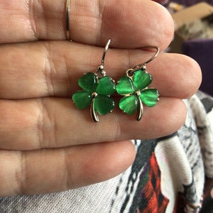May include: A pair of silver dangle earrings with green four-leaf clover charms. The clovers have a shiny, iridescent finish.