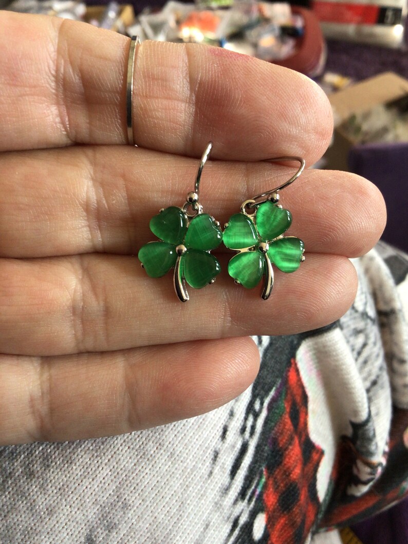 May include: A pair of silver dangle earrings with green four-leaf clover charms. The clovers have a shiny, iridescent finish.