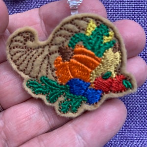 Puede incluir: Una cornucopia de fieltro marrón llena de verduras bordadas en tonos de naranja, rojo, verde y azul. La cornucopia está unida a una cadena plateada.