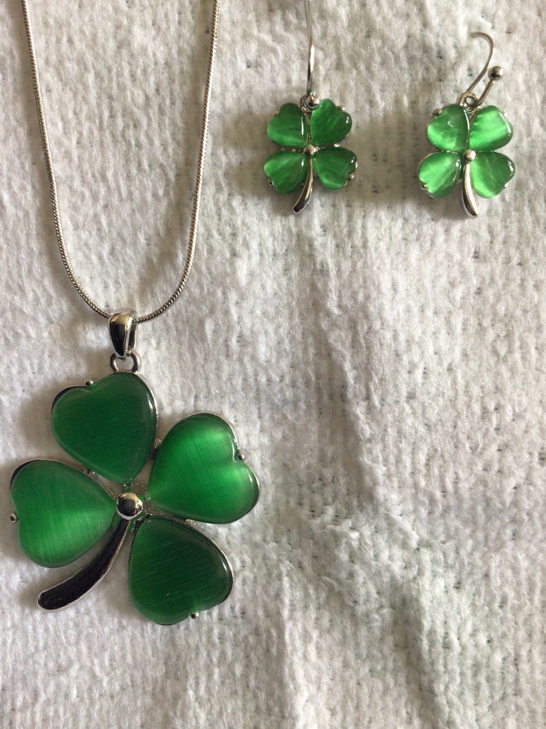 May include: A silver necklace and earrings set with green four-leaf clover charms. The charms have a shiny, iridescent finish.