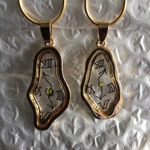 May include: Two gold chain necklaces with a melted clock pendant. The clock face is white with black Roman numerals and black hands. The pendant is clear and has a gold frame.