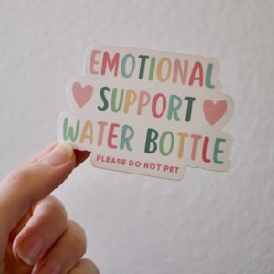 May include: A white sticker with the text "Emotional Support Water Bottle" in colourful letters. Two pink hearts are on either side of the word "Support". The text "Please do not pet" is printed in smaller letters below the main text.