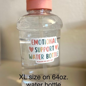 May include: A clear plastic water bottle with a pink lid and handle. The bottle has a sticker that says "Emotional Support Water Bottle" with hearts and the text "Please do not pet". The bottle is 64 fluid ounces and is labelled "XL size".