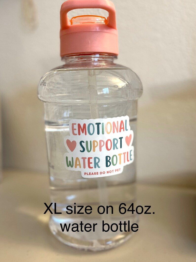 May include: A clear plastic water bottle with a pink lid and handle. The bottle has a sticker that says "Emotional Support Water Bottle" with hearts and the text "Please do not pet". The bottle is 64 fluid ounces and is labelled "XL size".