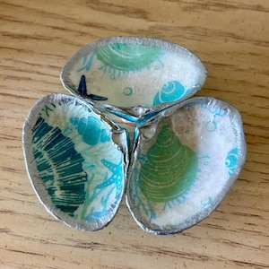 May include: A decorative dish crafted from three joined seashells, each featuring a decoupage pattern of teal and green sea life, including starfish and various shells, with the outer edges of the shells finished in a metallic silver paint.