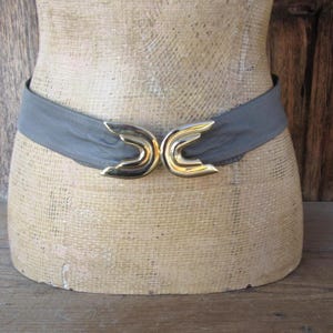 Puede incluir: Un cinturón gris con una hebilla dorada. La hebilla tiene un diseño único y el cinturón está envuelto alrededor de un maniquí. El cinturón es un accesorio de moda vintage.