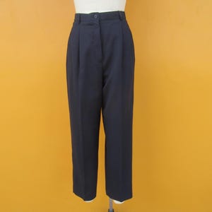 May include: Navy blue high-waisted trousers with a button closure and pleats. The pants are displayed on a mannequin against a yellow background. The trousers have a classic, tailored design.