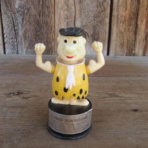 May include: A vintage Fred Flintstone push button puppet toy. The figure is wearing a yellow outfit with black spots and a black hat. The base of the toy has text that reads "FRED FLINTSTONE PUSH BUTTON PUPPET".