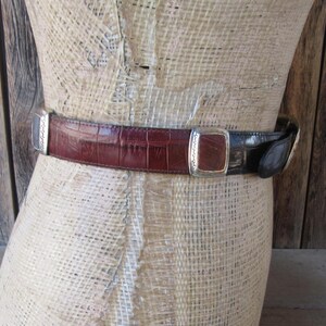 Pu&ograve; includere: Una cintura bicolore con un design in pelle goffrata a coccodrillo bordeaux e nera. La cintura presenta fibbie rettangolari argentate con bordi decorativi. La cintura &egrave; esposta su un manichino.