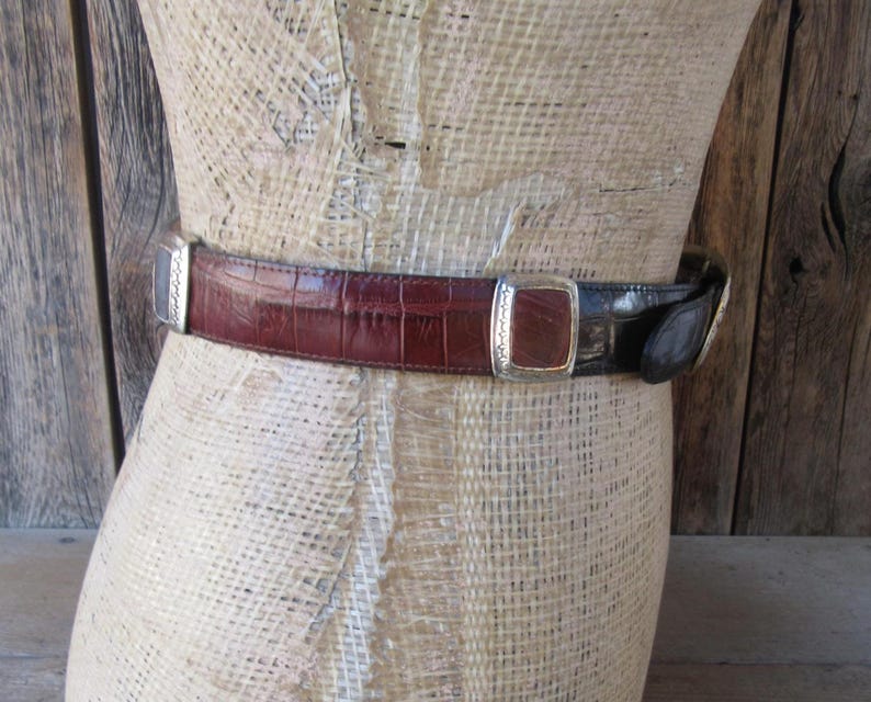 Pu&ograve; includere: Una cintura bicolore con un design in pelle goffrata a coccodrillo bordeaux e nera. La cintura presenta fibbie rettangolari argentate con bordi decorativi. La cintura &egrave; esposta su un manichino.