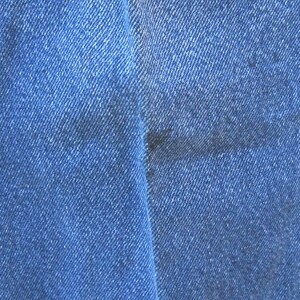 May include: Close-up of blue denim jeans fabric. The jeans have a classic design with a visible seam and a dark stain on the fabric. The denim has a textured appearance.