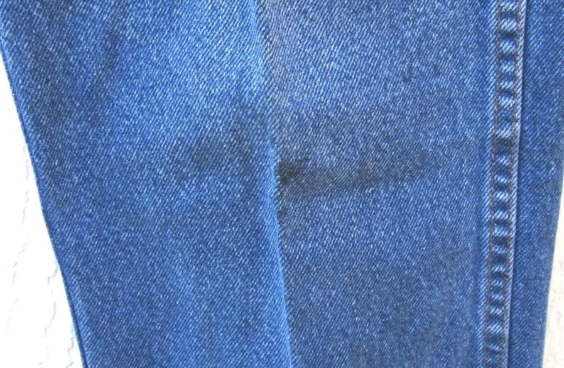 May include: Close-up of blue denim jeans fabric. The jeans have a classic design with a visible seam and a dark stain on the fabric. The denim has a textured appearance.