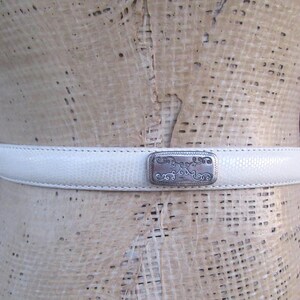 May include: A white belt with a textured surface and a silver-colored buckle. The buckle has an ornate design. The belt is displayed on a mannequin.