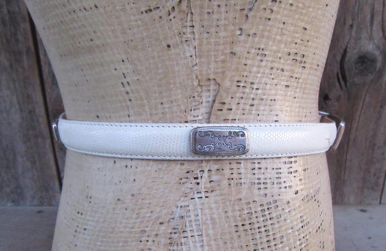 May include: A white belt with a textured surface and a silver-colored buckle. The buckle has an ornate design. The belt is displayed on a mannequin.