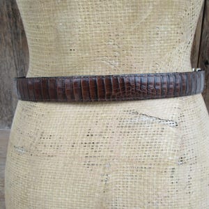 May include: A brown, textured belt with a reptile-like pattern. The belt is wrapped around a mannequin form. The belt has a silver-colored buckle. The background is a light tan color with wooden planks.