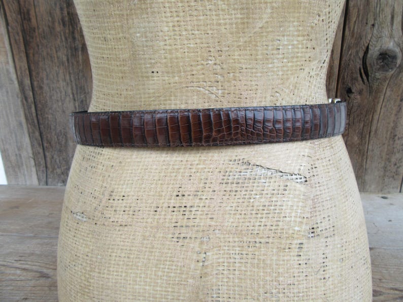 May include: A brown, textured belt with a reptile-like pattern. The belt is wrapped around a mannequin form. The belt has a silver-colored buckle. The background is a light tan color with wooden planks.