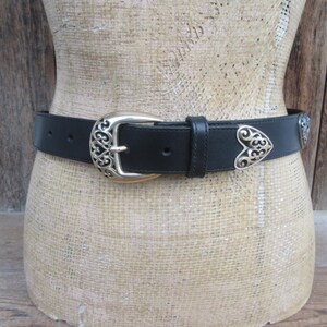 Op de afbeelding: Zwarte leren riem met een zilverkleurige gesp en hartvormige accenten. De gesp en accenten hebben een sierlijk, opengewerkt ontwerp. De riem wordt op een mannequin getoond.