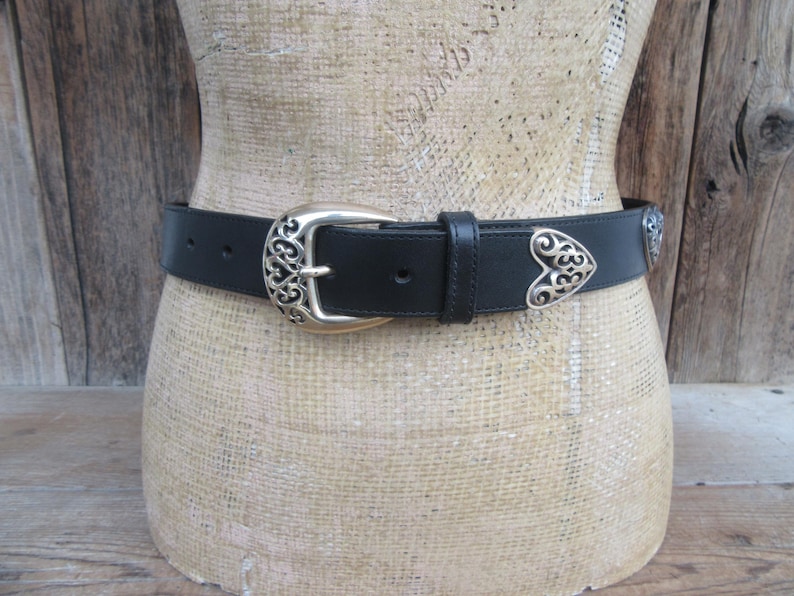 Op de afbeelding: Zwarte leren riem met een zilverkleurige gesp en hartvormige accenten. De gesp en accenten hebben een sierlijk, opengewerkt ontwerp. De riem wordt op een mannequin getoond.