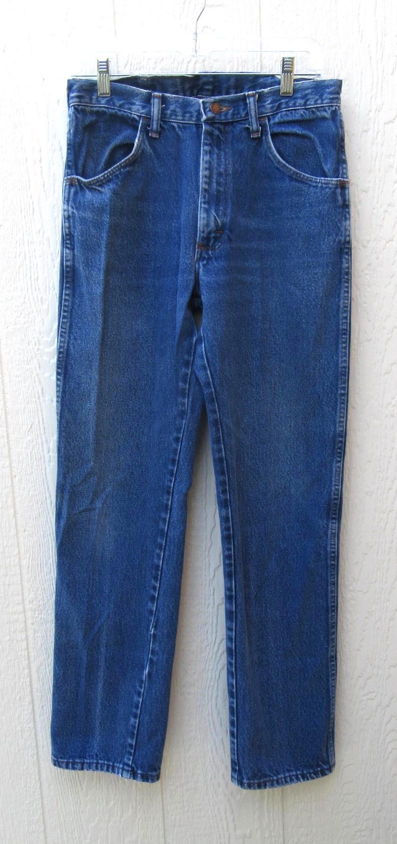 May include: Classic blue denim jeans with a straight leg cut. The jeans feature traditional five-pocket styling, a button and zipper closure, and belt loops. The garment is hanging on a metal hanger.