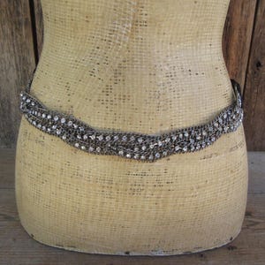 May include: A silver-toned belt with a woven design and sparkling rhinestone accents. The belt is displayed on a mannequin with a neutral-toned surface, set against a rustic wooden backdrop. The belt is a fashion accessory.