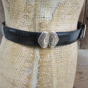 May include: Black belt with a textured, crocodile-like pattern. The belt features a decorative silver-toned buckle with an ornate design. The belt is displayed on a mannequin.