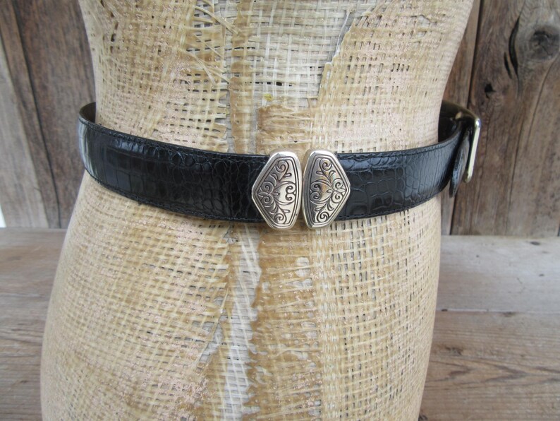 May include: Black belt with a textured, crocodile-like pattern. The belt features a decorative silver-toned buckle with an ornate design. The belt is displayed on a mannequin.