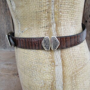 May include: A brown leather belt with a crocodile texture is fastened around a mannequin. The belt has a silver buckle and a decorative silver clasp in the center. The belt is a medium width.