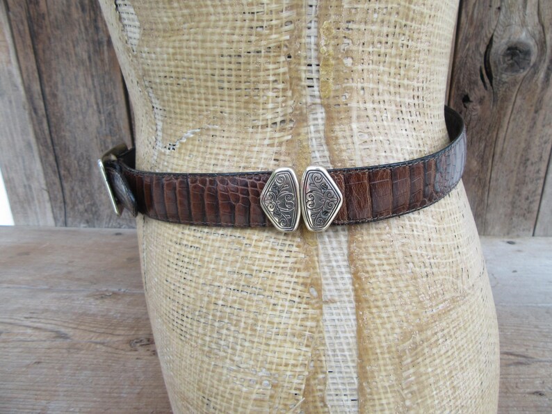 May include: A brown leather belt with a crocodile texture is fastened around a mannequin. The belt has a silver buckle and a decorative silver clasp in the center. The belt is a medium width.