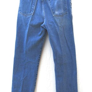 May include: Blue denim jeans with a classic five-pocket design. The jeans have a straight leg cut and are hanging from a hanger. The back pockets and waistband are visible. The jeans are made of a durable denim material.