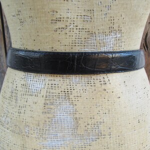 May include: A black belt with a textured, crocodile-like pattern is displayed on a mannequin. The belt is a solid black colour and appears to be made of leather or a similar material. The mannequin is a neutral colour, and the background is a wooden surface.