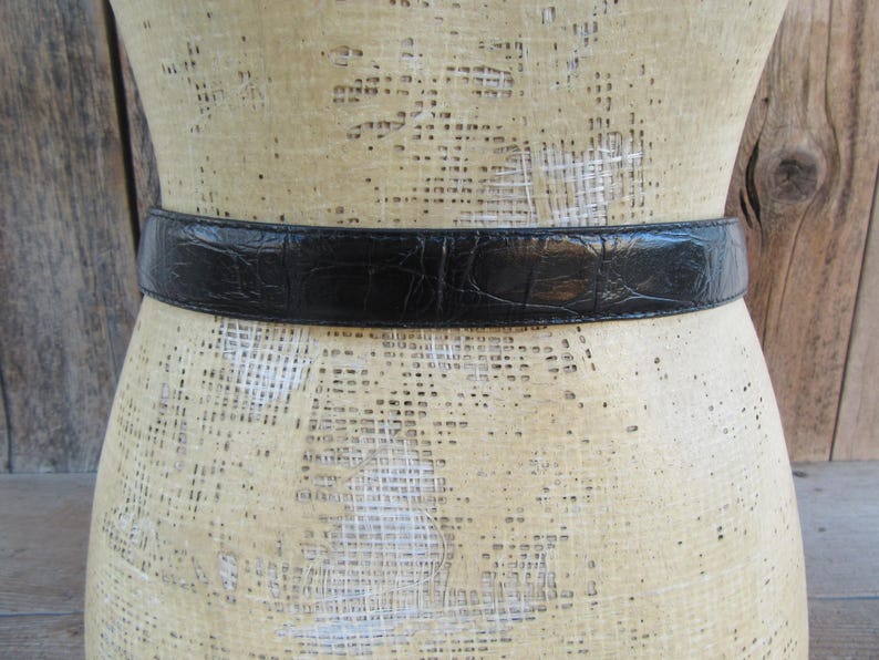 May include: A black belt with a textured, crocodile-like pattern is displayed on a mannequin. The belt is a solid black colour and appears to be made of leather or a similar material. The mannequin is a neutral colour, and the background is a wooden surface.