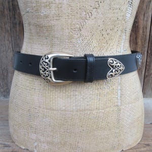 Op de afbeelding: Zwarte leren riem met een zilverkleurige gesp en hartvormige accenten. De gesp heeft een sierlijk ontwerp en de riem wordt op een mannequin getoond. Een klassiek accessoire.