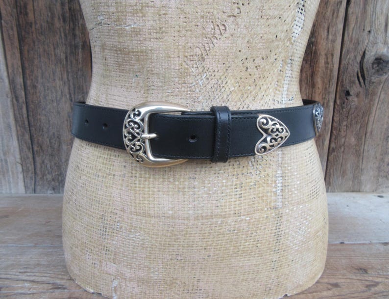 Op de afbeelding: Zwarte leren riem met een zilverkleurige gesp en hartvormige accenten. De gesp heeft een sierlijk ontwerp en de riem wordt op een mannequin getoond. Een klassiek accessoire.