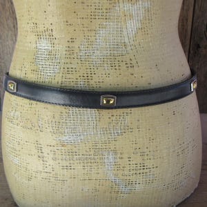 May include: A black leather belt with gold-toned square accents. The belt is worn around a mannequin. The gold accents are square with a gold center. The belt is a simple, classic design.