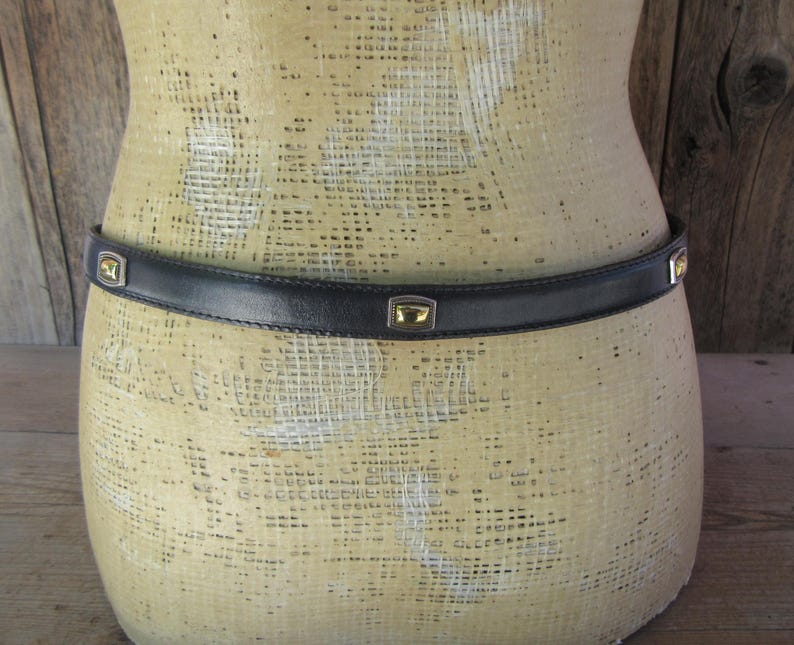 May include: A black leather belt with gold-toned square accents. The belt is worn around a mannequin. The gold accents are square with a gold center. The belt is a simple, classic design.