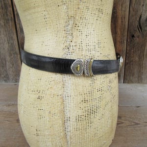 K&ouml;nnte beinhalten: Schwarzer Lederg&uuml;rtel mit dekorativer Silber- und Gold-Schnalle. Der G&uuml;rtel wird um eine Schaufensterpuppe getragen. Die Schnalle hat ein geometrisches Design mit einem goldfarbenen Zentralelement. Ein klassisches Accessoire.