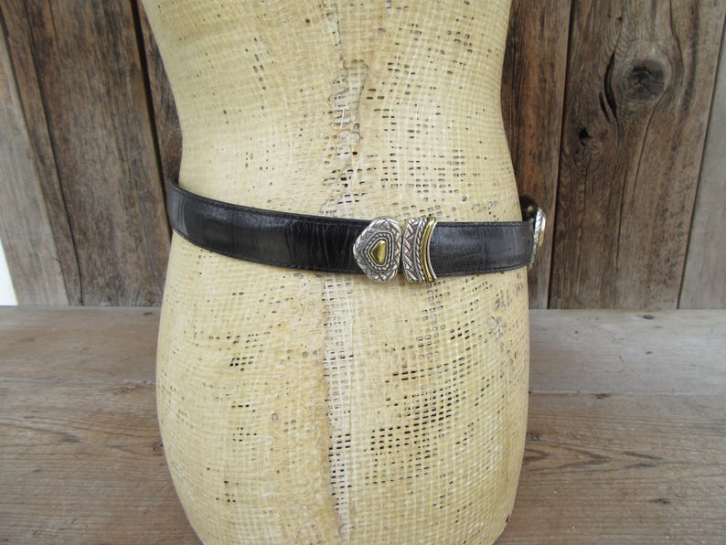 K&ouml;nnte beinhalten: Schwarzer Lederg&uuml;rtel mit dekorativer Silber- und Gold-Schnalle. Der G&uuml;rtel wird um eine Schaufensterpuppe getragen. Die Schnalle hat ein geometrisches Design mit einem goldfarbenen Zentralelement. Ein klassisches Accessoire.